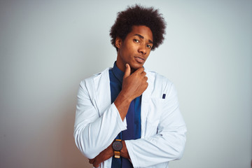 Wall Mural - Young african american doctor man wearing coat standing over isolated white background looking confident at the camera smiling with crossed arms and hand raised on chin. Thinking positive.