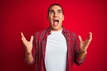 Canvas Print - Young handsome man wearing a shirt using watch over red isolated background crazy and mad shouting and yelling with aggressive expression and arms raised. Frustration concept.