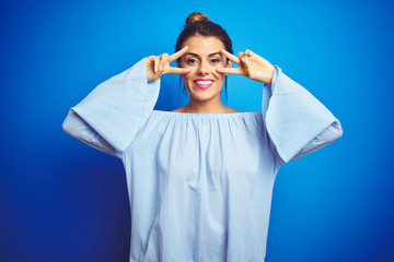 Sticker - Young beautiful woman wearing bun hairstyle over blue isolated background Doing peace symbol with fingers over face, smiling cheerful showing victory