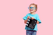 Funny smiling child girl in cinema glasses hold film making clapperboard isolated on pink background. Studio portrait. Childhood lifestyle concept. Copy space for text.