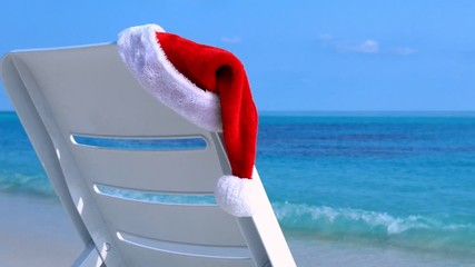 Wall Mural - Santa Claus Hat on sunbed near calm beach with azure sea water and white sand. Christmas vacation on island