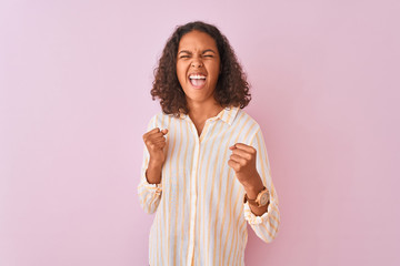 Wall Mural - Young brazilian woman wearing striped shirt standing over isolated pink background very happy and excited doing winner gesture with arms raised, smiling and screaming for success. Celebration concept.