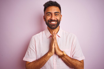 Canvas Print - Young indian man wearing casual shirt standing over isolated pink background praying with hands together asking for forgiveness smiling confident.