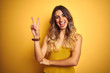 Young beautiful woman wearing t-shirt over yellow isolated background smiling with happy face winking at the camera doing victory sign with fingers. Number two.