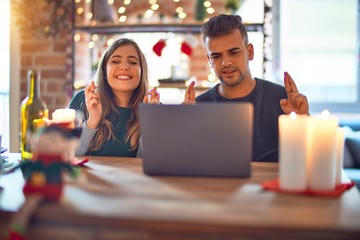 Young beautiful couple sitting using laptop around christmas decoration at home gesturing finger crossed smiling with hope and eyes closed. Luck and superstitious concept.