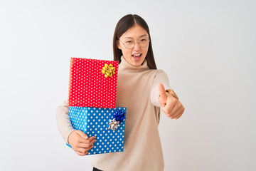 Canvas Print - Young chinese woman holding birthday gifts over isolated white background approving doing positive gesture with hand, thumbs up smiling and happy for success. Winner gesture.