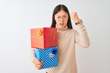 Sticker - Young chinese woman holding birthday gifts over isolated white background angry and mad raising fist frustrated and furious while shouting with anger. Rage and aggressive concept.