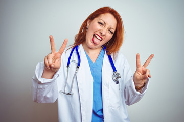 Poster - Young redhead doctor woman using stethoscope over white isolated background smiling with tongue out showing fingers of both hands doing victory sign. Number two.