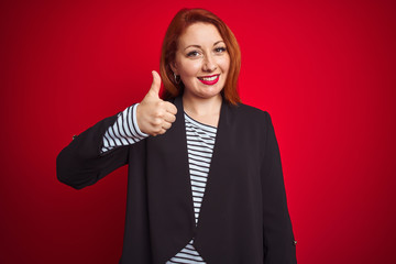 Poster - Beautiful redhead business woman wearing elegant jacket over isolated red background doing happy thumbs up gesture with hand. Approving expression looking at the camera with showing success.