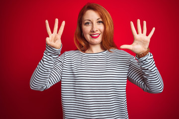 Wall Mural - Young redhead woman wearing strapes navy shirt standing over red isolated background showing and pointing up with fingers number eight while smiling confident and happy.