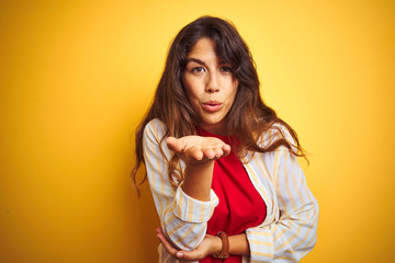 Wall Mural - Young beautiful woman wearing red t-shirt and stripes shirt over yellow isolated background looking at the camera blowing a kiss with hand on air being lovely and sexy. Love expression.