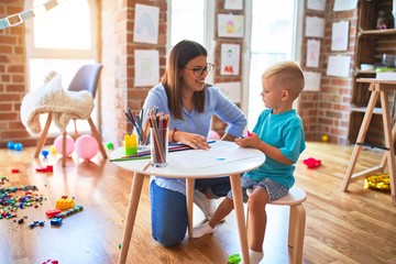 Young caucasian child playing at playschool with teacher. Mother and son at playroom drawing a draw with color pencils