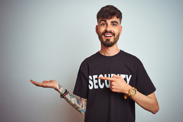 Sticker - Young safeguard man with tattoo wering security uniform over isolated white background amazed and smiling to the camera while presenting with hand and pointing with finger.