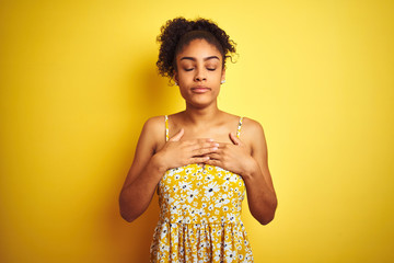 Sticker - African american woman wearing casual floral dress standing over isolated yellow background smiling with hands on chest with closed eyes and grateful gesture on face. Health concept.