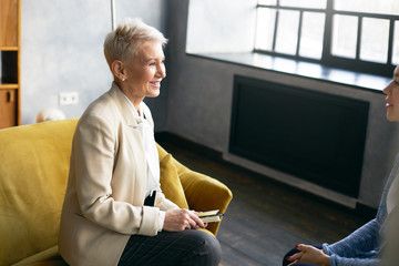 High angle view of beautiful elegant middle aged woman in beige suit sitting in armchair at her office and smiling encouraging female client during counselling. People, age, job and occupation concept