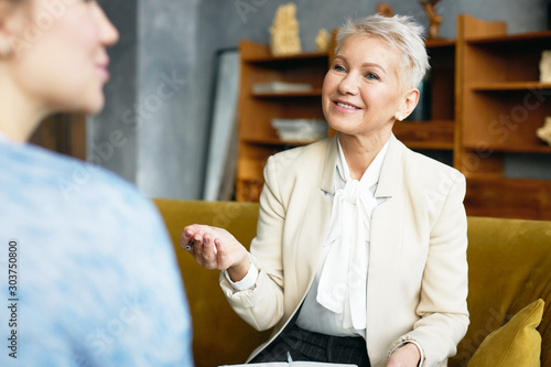 Cuadros en Lienzo Indoor shot of positive mature female psychologist in elegant suit sitting on couch and making notes while having therapy with unrecognizable woman patient
