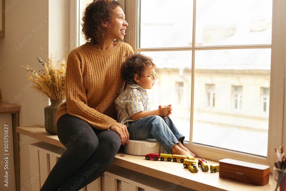 Cozy scene of joyful young Latin female wearing jeans and knitted jumper sitting on windowsill hugging her cute chubby little child, both watching outside, talking, having dreamy thoughtful expression - obrazy, fototapety, plakaty 