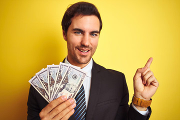 Wall Mural - Young handsome businessman wearing suit holding dollars over isolated yellow background very happy pointing with hand and finger to the side