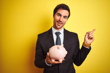 Canvas Print - Young handsome businessman holding piggy bank over isolated yellow background very happy pointing with hand and finger to the side