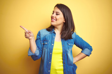 Sticker - Young beautiful brunette woman wearing casual denim jacket over yellow isolated background with a big smile on face, pointing with hand and finger to the side looking at the camera.