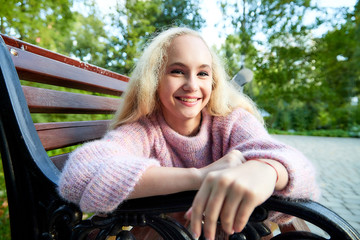 Pretty teenage girl 14-16 year old with curly long blonde hair in the green park on the bench in a summer day outdoors. Beautiful portrait