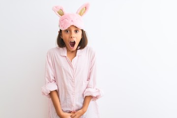 Poster - Beautiful child girl wearing sleep mask and pajama standing over isolated white background afraid and shocked with surprise expression, fear and excited face.