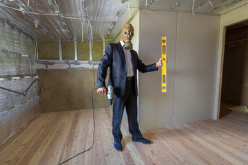  Strange man in businessman suit and gas protection mask inside a room under renovation works holding electric screwdriver and a level tools.