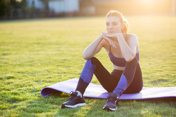 Wall Mural - Young sportive woman in sports clothes sitting on training mat before doing exercises in field at sunrise.