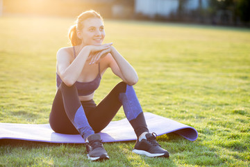 Young sportive woman in sports clothes sitting on training mat before doing exercises in field at sunrise.