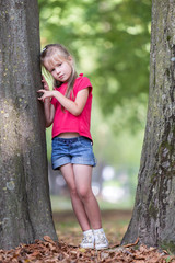 Canvas Print - Portrait of a pretty little child girl standing near big tree trunk in summer park outdoors.