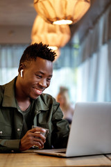 Wall Mural - Cheerful young african american student man in headphones using computer in cafe. Online video chat with friends