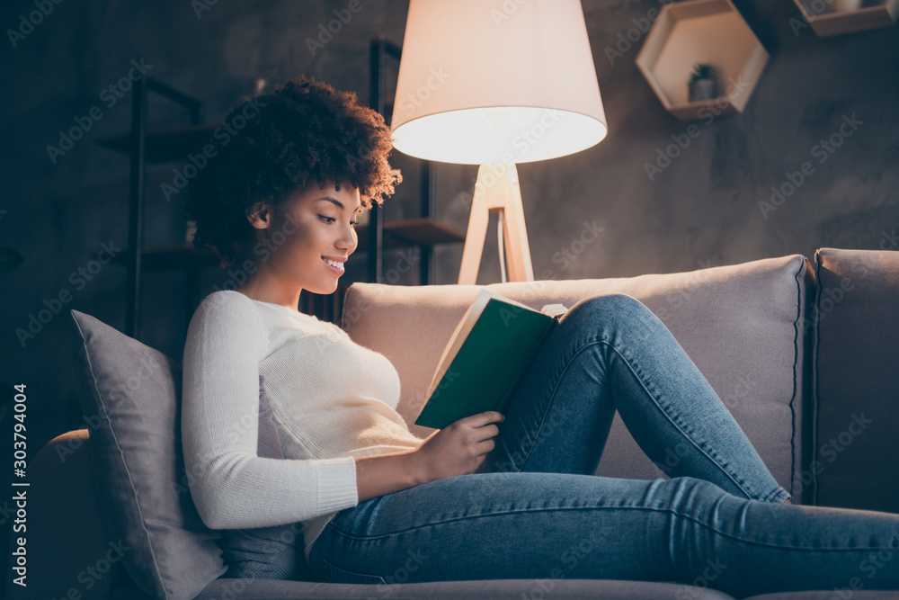 Photo of attractive dark skin curly lady holding favorite historic novel reading excited lying cozy couch wear casual sweater jeans outfit evening living room indoors - obrazy, fototapety, plakaty 