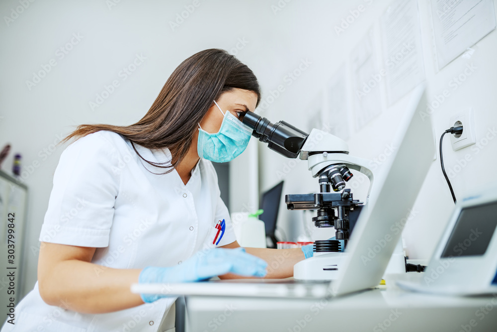 Charming caucasian laboratory assistant in white uniform, with protective mask and rubber gloves sitting in lab looking blood sample trough microscope and using laptop. - obrazy, fototapety, plakaty 