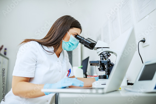 Cuadros en Lienzo Charming caucasian laboratory assistant in white uniform, with protective mask and rubber gloves sitting in lab looking blood sample trough microscope and using laptop