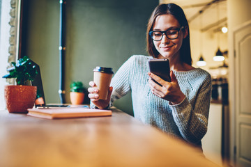 Cheerful hipster blogger with coffee to go in hands reading incoming sms message on smartphone connected to free wifi.Positive young student watching broadcasting online on modern mobile phone