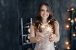 Girl with a sparkler near the Christmas tree. A cheerful young woman with a cute smile in a beige dress is standing and holding a sparkling sparkler in the hands against the background of the Christma