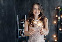 Girl With A Sparkler Near The Christmas Tree. A Cheerful Young Woman With A Cute Smile In A Beige Dress Is Standing And Holding A Sparkling Sparkler In The Hands Against The Background Of The Christma