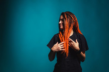 Girl With Red Fire Dreadlocks On Sea Wave Background Color. A Young Woman With Orange Dreadlocks And Piercings On A Blue Background. Beautiful Daring Informal Image Of A Girl With Red Yellow Dreadlock