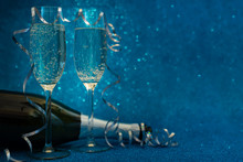 Two Glasses Of Champagne And Bottle Decorated Streamers On Blue Glittering Background. Christmas And New Year Holidays Concept. Copy Space.