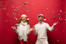 Excited Couple In Santa Hats Screaming And Celebrating With Golden Confetti, Isolated On Red