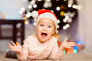 Wall Mural - Happy emotional little girl in Santa hat lying on floor at home