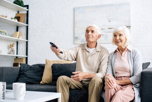 Fotomural smiling husband and wife watching tv and sitting on sofa in apartment