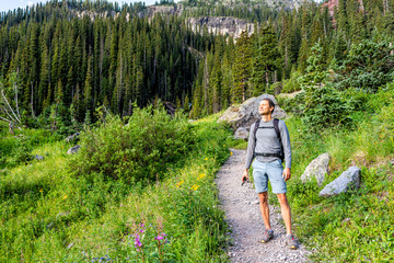 man hiker standing with backpack on green alpine rocky meadow by pink fireweed wildflowers on trail 