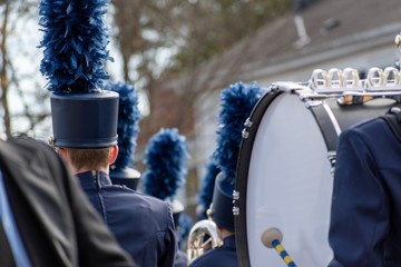 A high school marching band performs during a Veterans Day parade. Photo taken from the back and displays a french horn and bass drum player with traditional uniform. Taken in landscape orientation.