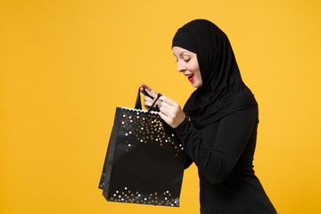 Arabian muslim woman in black hijab hold package bag, purchases after shopping isolated on yellow background studio portrait. Birthday holiday people religious lifestyle concept. Mock up copy space.