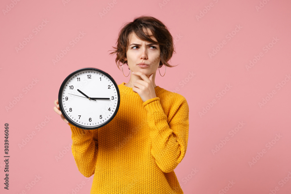 Pensive young brunette woman girl in yellow sweater posing isolated on pastel pink background, studio portrait. People lifestyle concept. Mock up copy space. Holding clock, put hand prop up on chin. - obrazy, fototapety, plakaty 