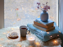 Cozy Home. Winter Holidays Still Life. Cup Of Tea With Sweet Dessert, Candle, Books, Flowers In Blue Vase And Garland Lights At Window. Relaxing At Cold Season At Home. Copy Space