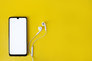 Smartphone with blank white screen connects to earphones on yellow background - top view. Earphone connected to cellphone.