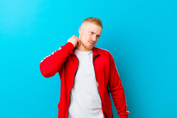 young blonde man wearing sports clothes feeling stressed, frustrated and tired, rubbing painful neck, with a worried, troubled look against blue background