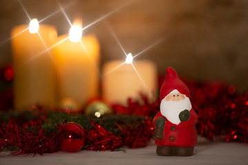 The warmth of the Christmas concept: a Santa Claus statuette in foreground in a rustic setting with with three candles lit in bokeh effect, gold and red bright baubles, cross screen star effect